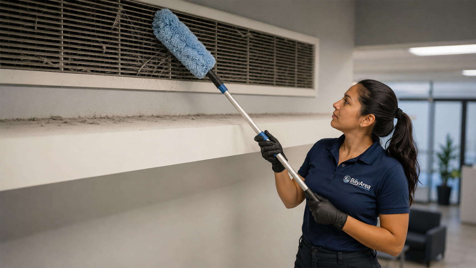 Professional Bay Area Cleaners technician performing high dusting in an office, removing dust and cobwebs from an air vent and upper ledge with a microfiber extension tool.